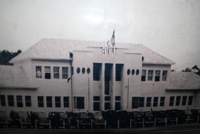Na imagem em preto e branco há a presença de um céu nublado à luz do dia, algumas àrvores ao fundo e a fachada da escola ao centro, na cor branca.