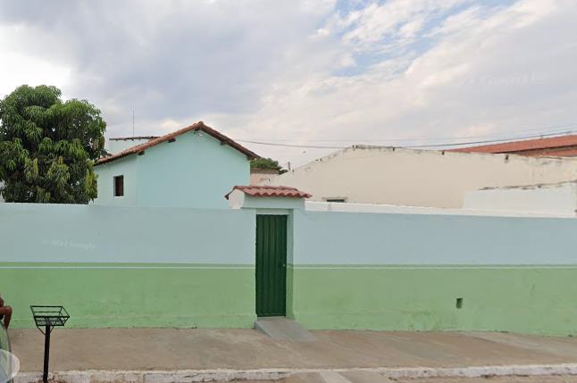 A imagem apresenta um grande muro nas cores azul e verde claro com um portão estreito no meio na cor verde escuro. Atrás há um telhado com uma janela e uma parte de uma árvore no lado esquerdo. O céu está carregado de nuvens.