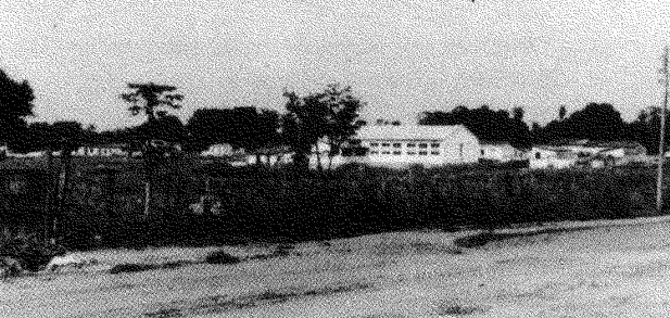 É uma fotografia em preto e branco de uma escola com diversas janelas pequenas. Ao lado tem árvores, mato e uma possível estrada de terra.