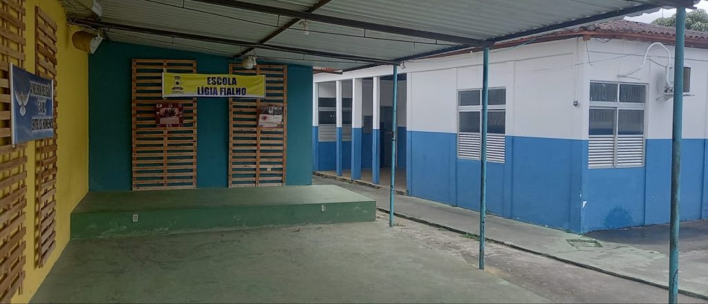 Na imagem há uma escola em um espaço com grades e paletes na parede com fundo amarelo do lado esquerdo. Ao lado direito há salas de aula com janela nas cores branco e azul. 