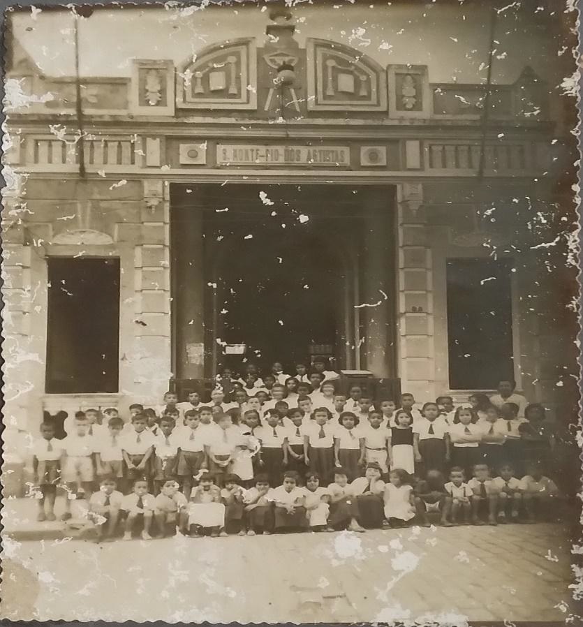 A imagem está em preto e branco com um prédio com corredor central e uma janela de cada lado. À frente há crianças uniformizadas de branco e preto com gravatas.