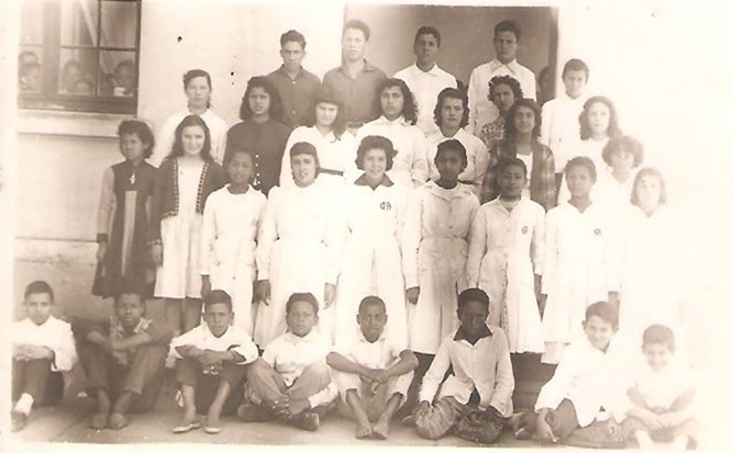 A imagem retrata uma fotografia antiga em preto e branco com crianças e adolescentes vestidos de branco, prováveis aluno e adultos como professores e professoras.