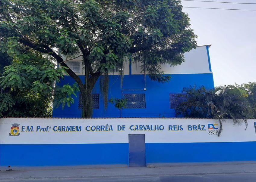 A imagem apresenta a fachada da escola com um prédio nas cores azul e branco e um portão azul escuro no meio. Em cima está escrito ESCOLA MUNICIPAL PROFESSORA CARMEM CORRÊA DE CARVALHO REIS BRÁZ. E também há uma árvore logo atrás do muro.