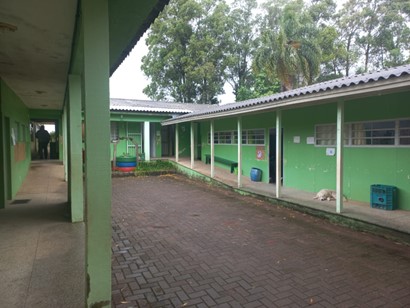 A imagem apresenta uma escola pintada nas cores verde bandeira e branco. Tem um pátio no meio da estrutura com diversas salas em um corredor.