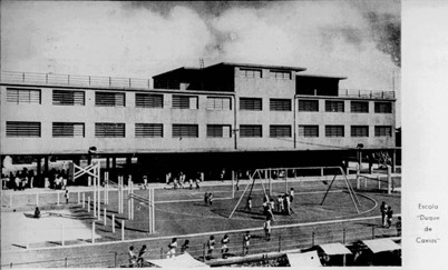 A imagem está em preto e branco. Há um enorme galpão com diversas janelas, à frente tem uma quadra de esportes com crianças brincando.