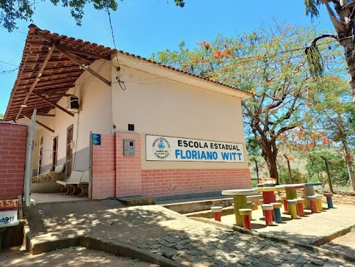 A imagem mostra a entrada da escola, por onde os alunos passam por uma rampa e portão. O muro da escola é de tijolinhos vermelhos, e uma parte é pintada de rosa, com uma placa com o nome da escola. Na frente da placa possui três mesas com banquinhos.