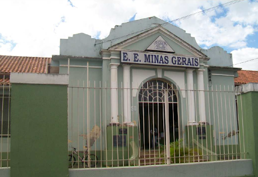 Imagem da fachada do Grupo Escolar de Minas Gerais, a fachada está centralizada na imagem e possui tons de verde e branco, com uma grande placa com o nome da escola.