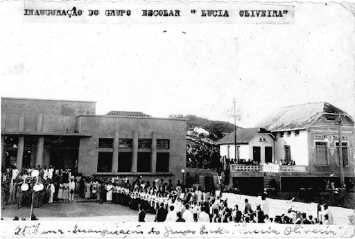 A foto monstra a inauguração da escola, com muitas pessoas em frete a entrada e poste de iluminação na calçada. Ao fundo está o prédio da escola, no canto esquerdo e uma casa no canto direito.