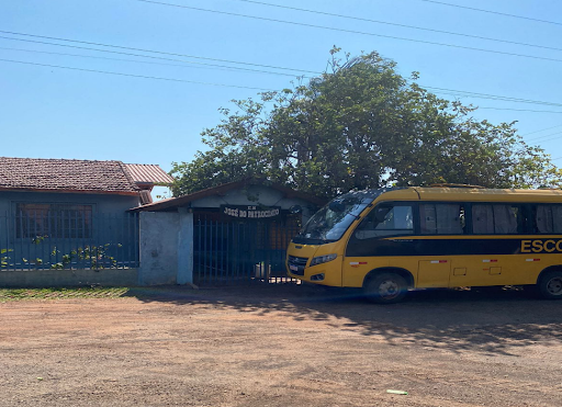 Na imagem um ônibus amarelo escolar aparece na frente da construção da Escola Municipal José do Patrocínio. À frente, o chão sem asfalto ocupa metade da imagem que tem, ao fundo, uma grande árvore verde. 