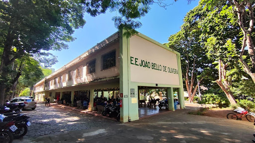 Fotografia lateral da Escola Estadual João Belo de Oliveira. Na imagem aparecem motocicletas, carros estacionados, pilotis ocupados por pessoas reunidas e um prédio em verde claro e amarelo que abriga a escola. 