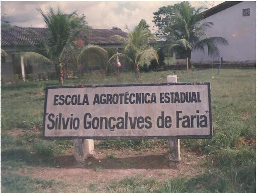 A imagem mostra a placa com o nome da Escola Agrotécnica Estadual Sílvio Gonçalves de Faria. Ao fundo, coqueiros ainda pequenos e um longo telhado ocupa o ponto de fuga da imagem. 