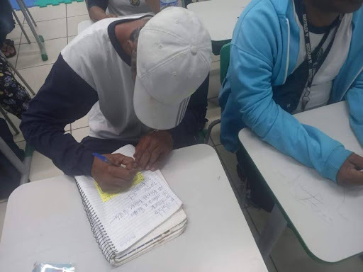 Na imagem aprecem dois estudantes em sala de aula. O primeiro escrevendo algo em seu caderno, de boné cinza claro e uniforme branco. O segundo, de blusa de frio azul, repousa o braço sobre a mesa. 